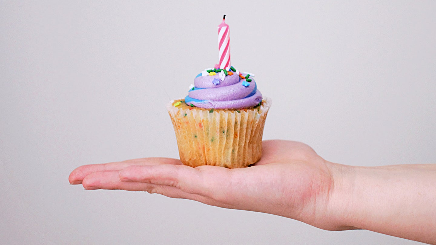 Hand holding a cupcake with purple frosting, sprinkles and a birthday candle.