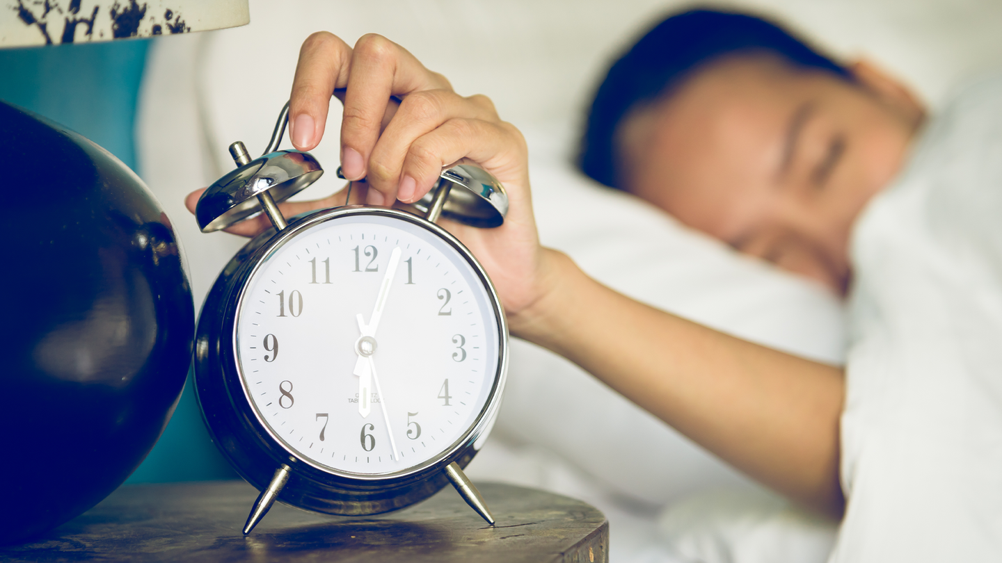 A woman just waking up in bed turns off an analog alarm clock.